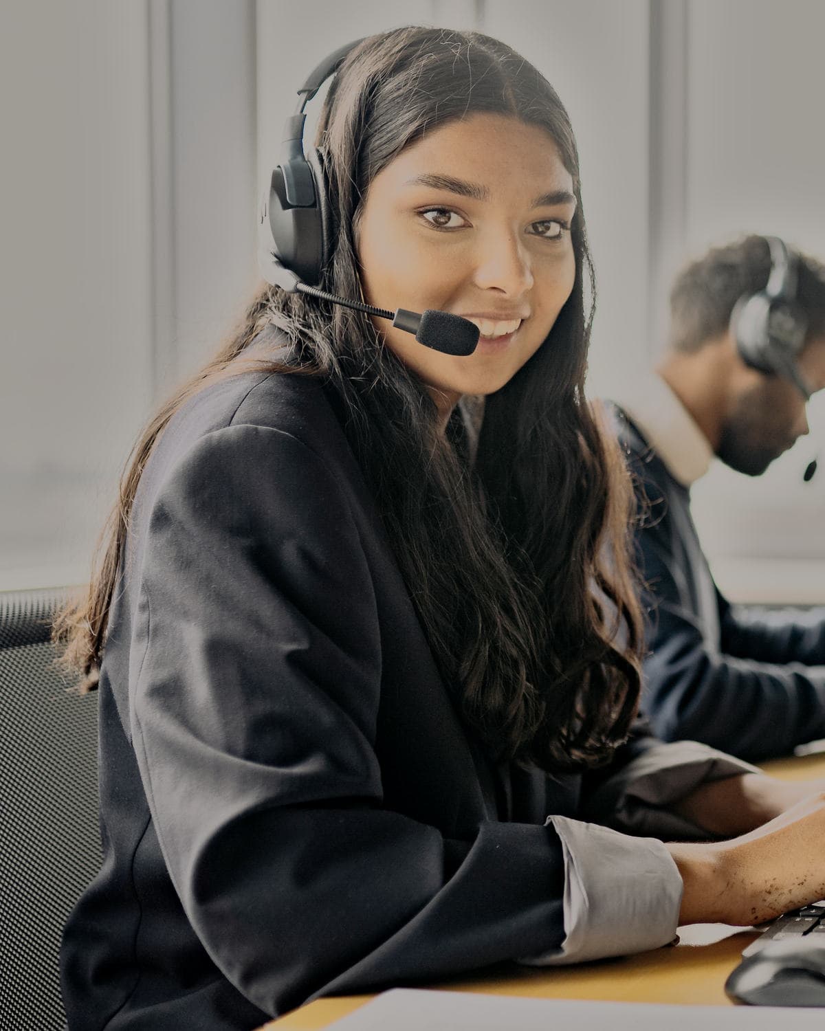 A woman in a headset smiles over her shoulder from her desk, a colleague at the next one