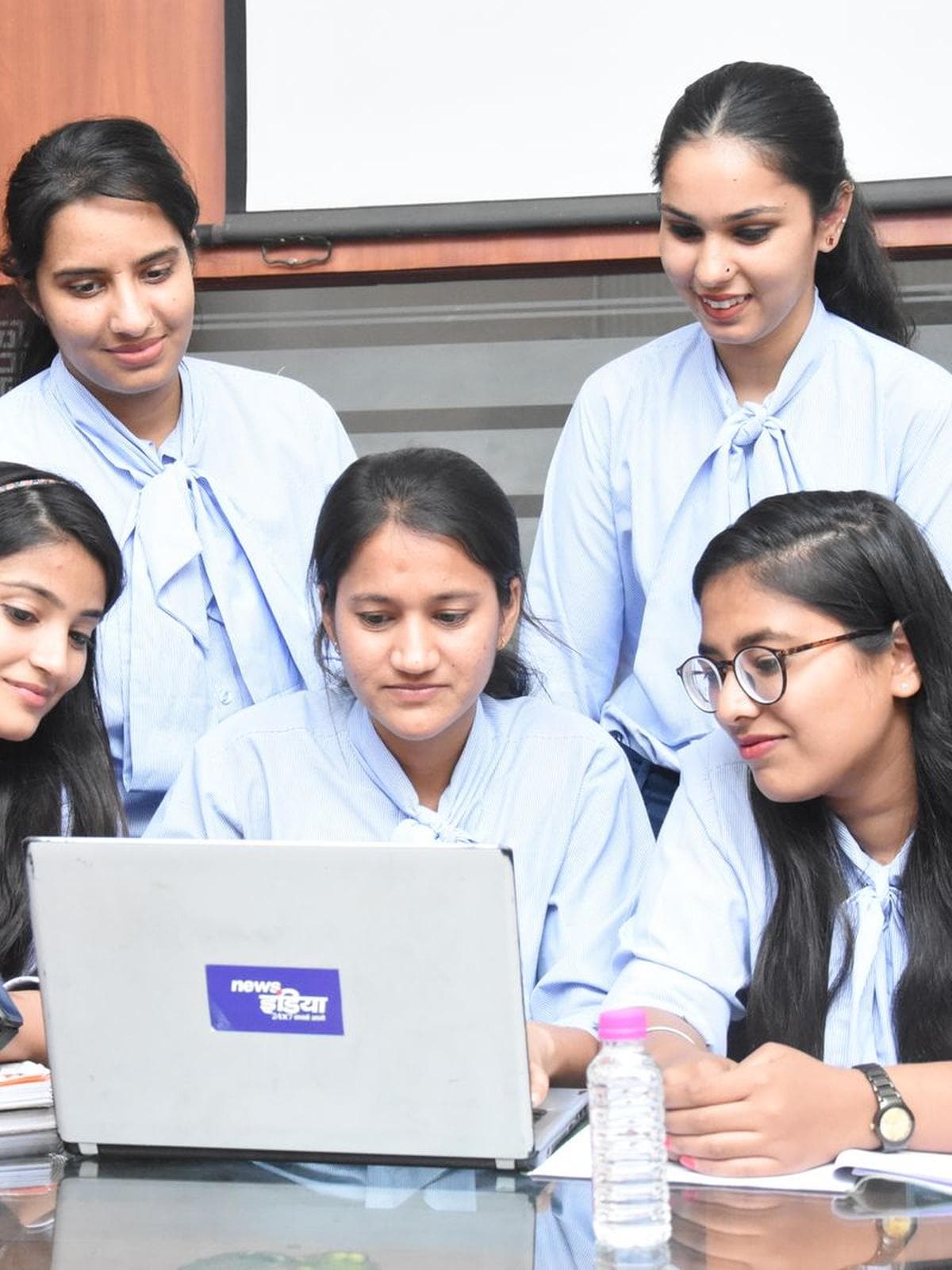 Five South Asian students in pale uniforms gathered around a laptop in a classroom, working through something together