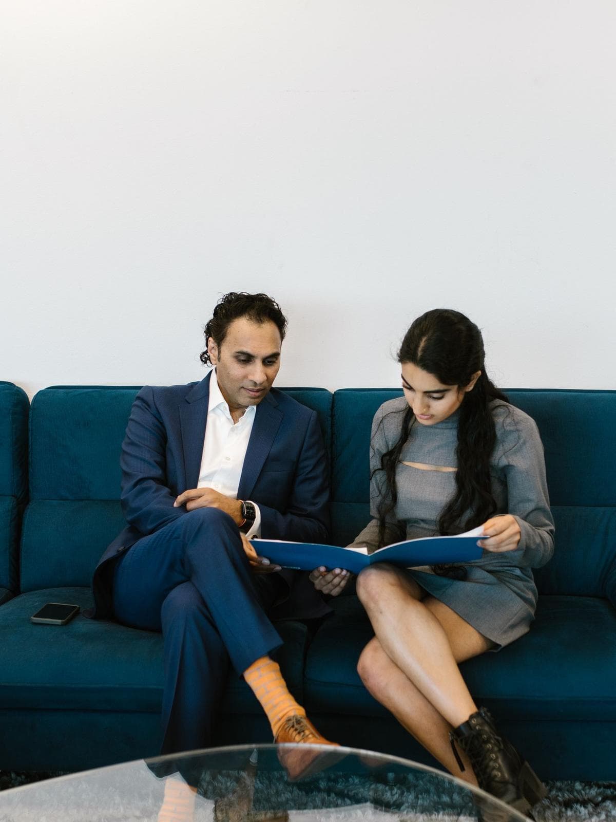 A South Asian advisor in a navy suit sitting on a blue sofa beside a student, the two of them reading through an open folder
