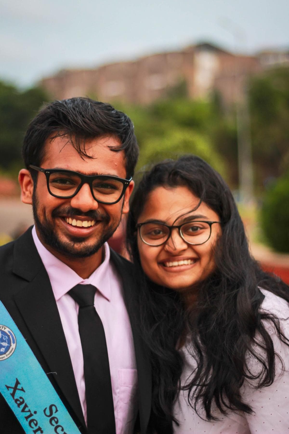 Two South Asian graduates in glasses, one in a suit with a sash and one in a pale sweater, standing together outdoors and smiling
