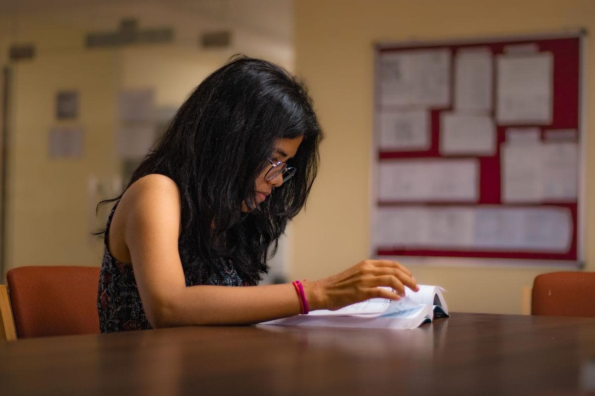 A South Asian student in glasses working through papers at a wooden desk in a quiet, warmly lit study room