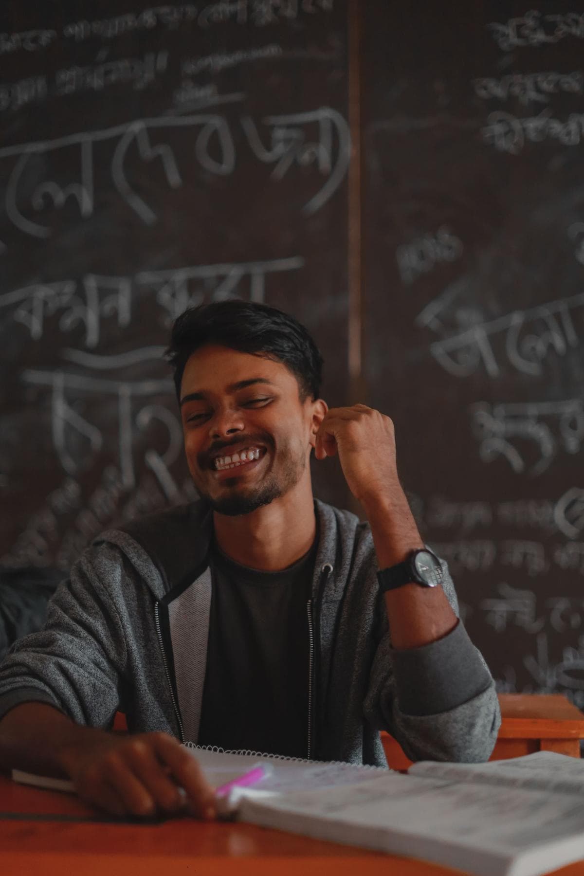 A smiling young Bangladeshi student at a desk in front of a blackboard covered in Bengali handwriting