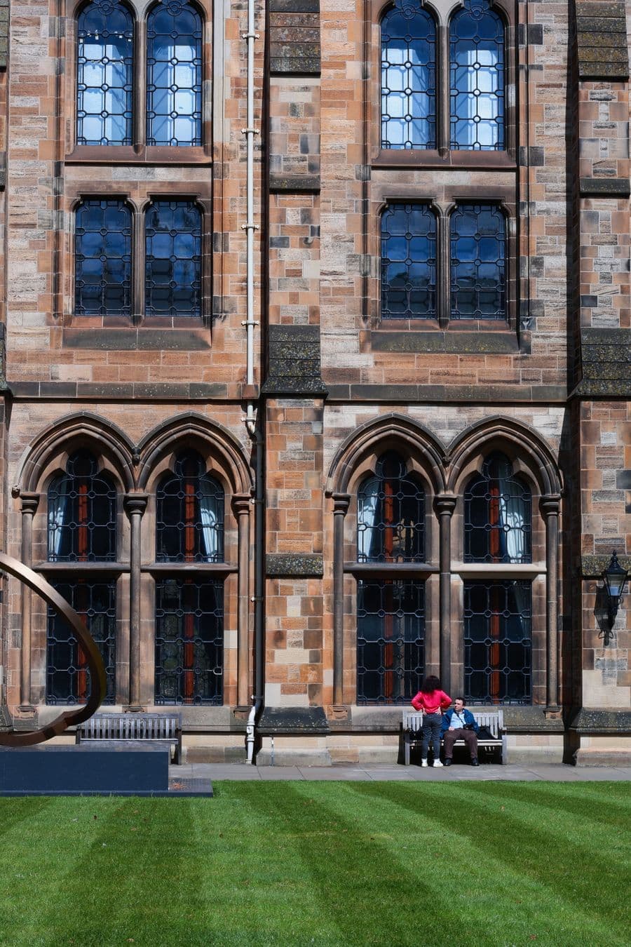 Ornate arched windows on a Gothic university facade, with visitors seated on a bench below