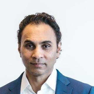A South Asian man with dark curly hair in a navy blazer and open white shirt, looking calmly at the camera against a plain white wall.