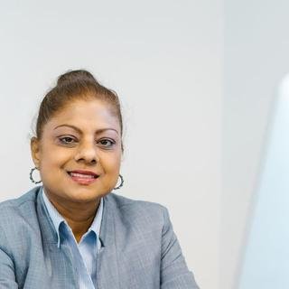 A smiling South Asian woman with her hair tied up, in a grey blazer and hoop earrings, seated at a bright office desk.