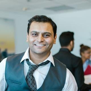 A smiling South Asian man in a teal waistcoat, shirt, and patterned tie, standing in an open-plan office.