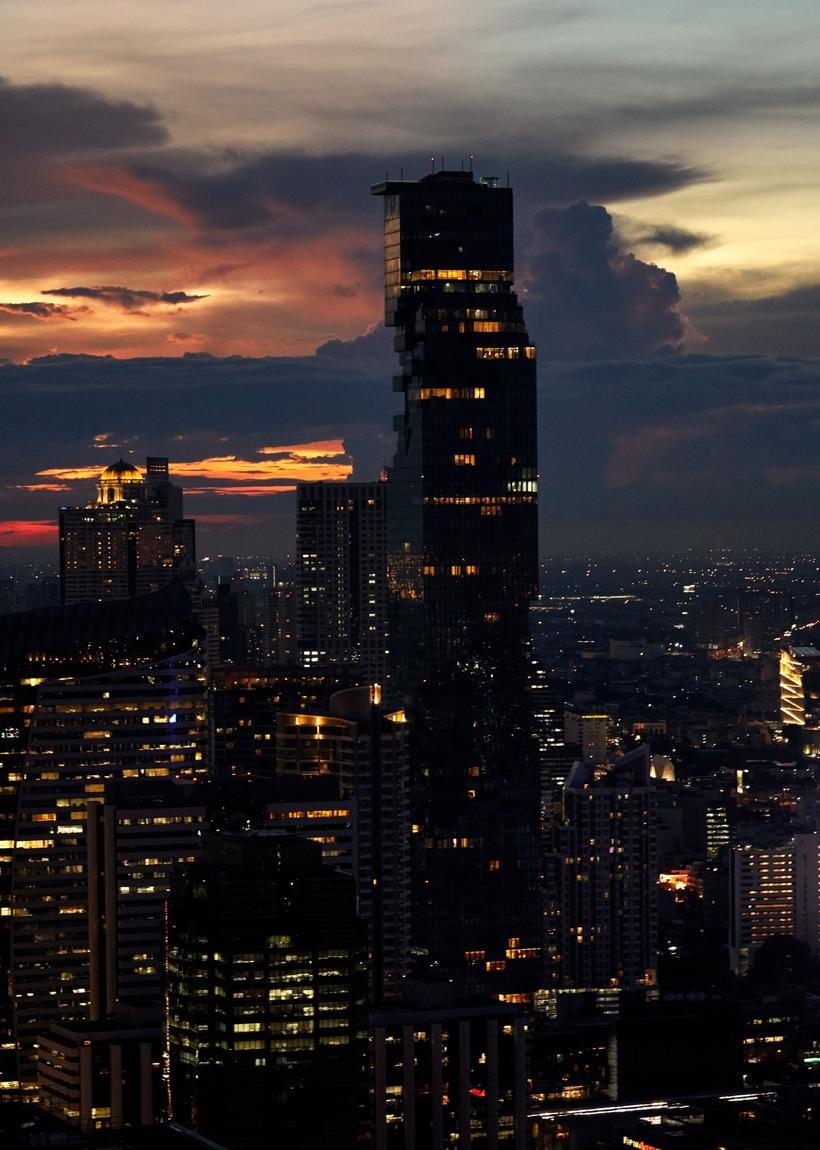 The Bangkok skyline at twilight, tall dark towers stacked across the frame under a burning orange and grey cloud bank.