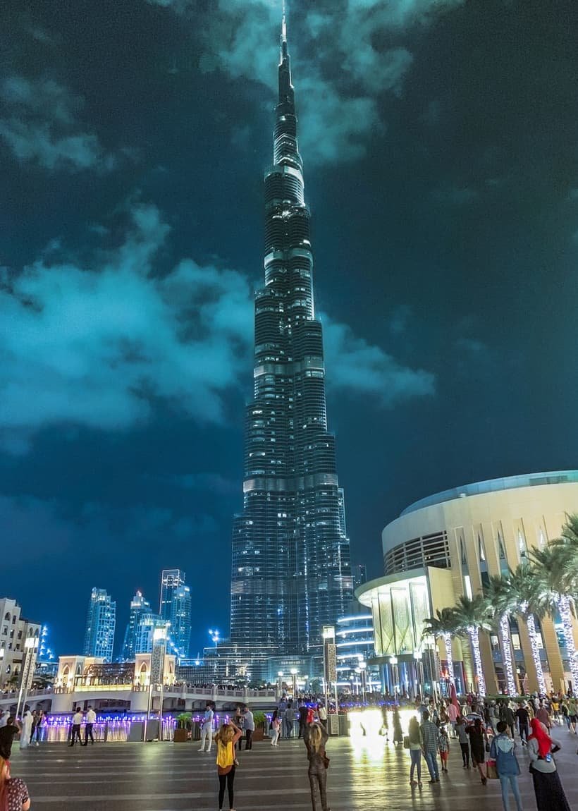 The Burj Khalifa tower rising far above a bright downtown Dubai plaza at night, with palm trees, lit shopfronts, and crowds of people below.