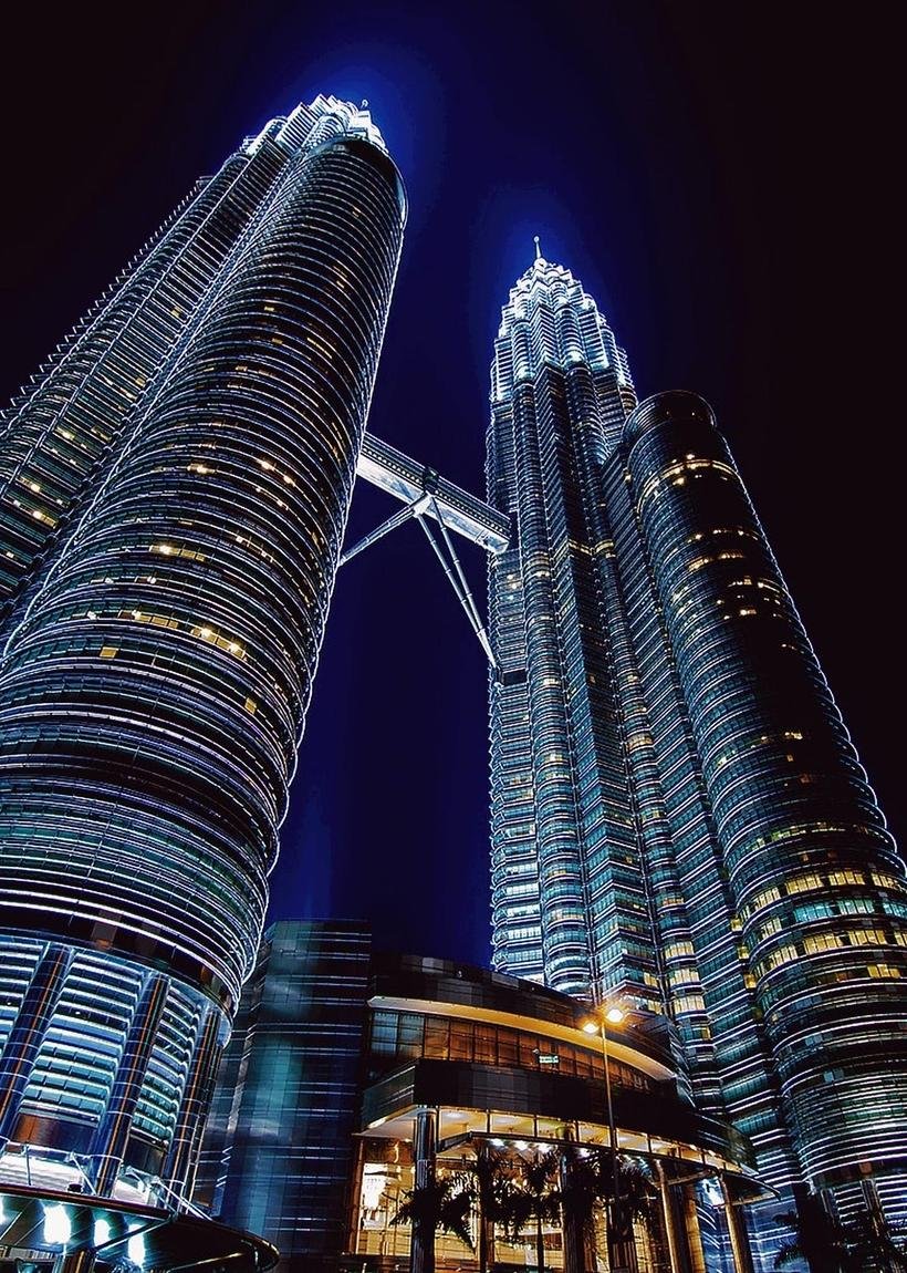 The Petronas Twin Towers in Kuala Lumpur seen from below at night, their steel facades and the sky bridge lit white against a deep blue sky.
