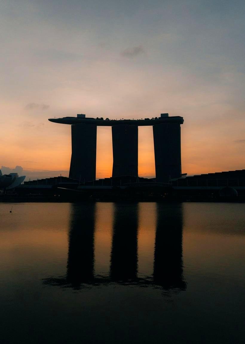 Marina Bay Sands in Singapore silhouetted against a burning orange dusk sky, its three towers and the long boat-shaped roof deck mirrored in the still water of the bay, the lotus-shaped ArtScience Museum at the left edge.