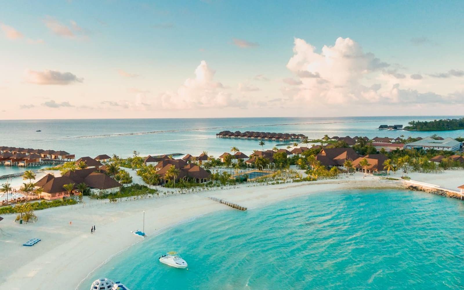 An aerial view of a tropical island resort at dusk, rows of thatched villas along a curving white-sand beach with calm turquoise water in the foreground and a soft pink sky.