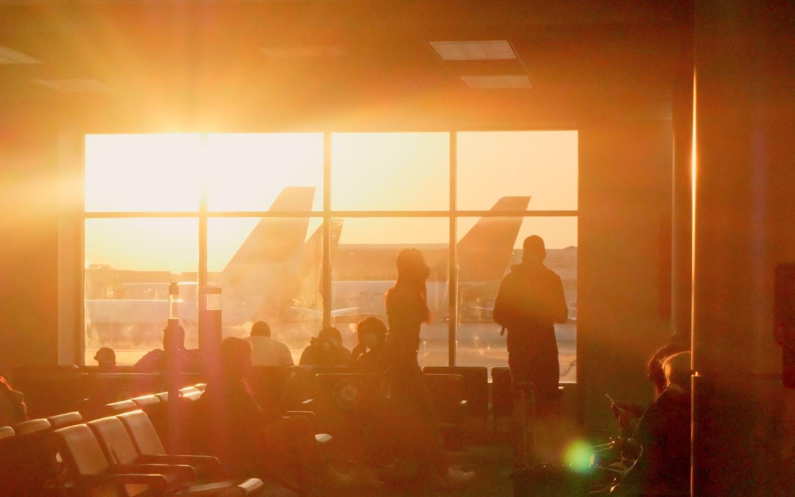 Travellers waiting at a departure gate at sunrise, in silhouette against a window wall with the tails of two aircraft beyond it.