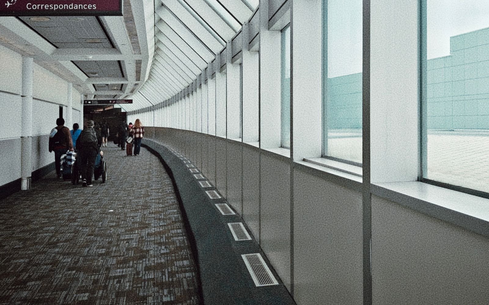 A landscape view of a gently curving, daylit airport corridor, travellers walking away in the distance beside a full-height window wall, a maroon overhead sign reading Correspondances.