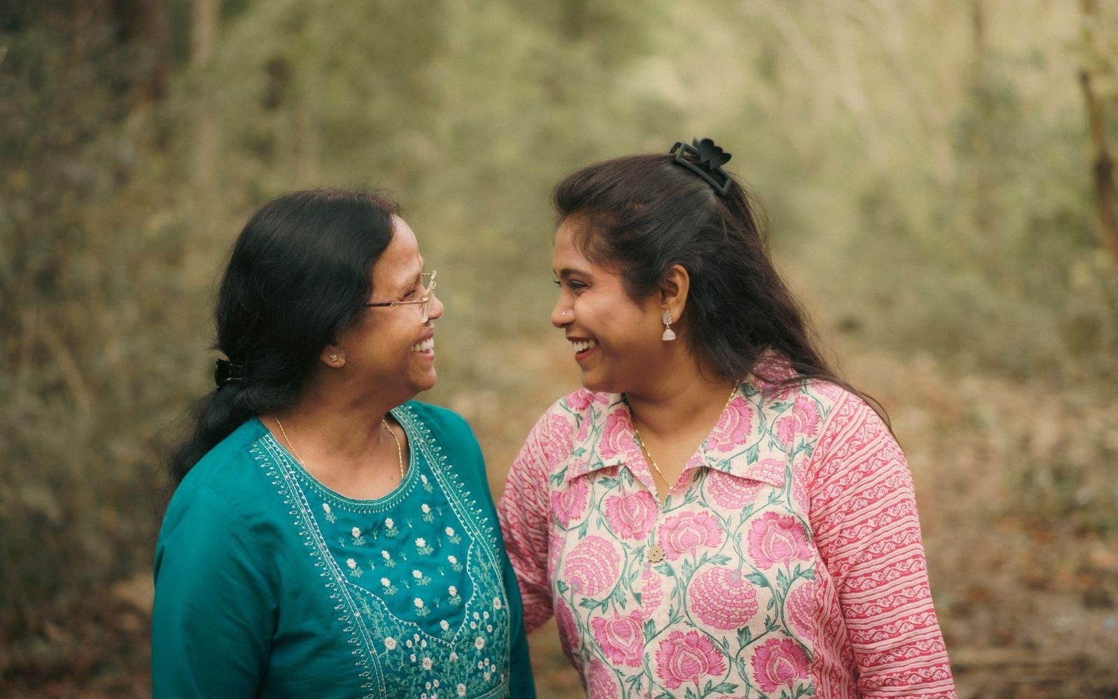 Two South Asian women, an older and a younger, stand close together outdoors in soft daylight, both in embroidered kurtas, smiling at each other mid-conversation.