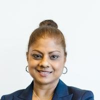 A smiling South Asian woman in a navy blazer with her hair tied up, photographed against a plain white wall.