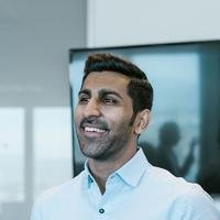 A smiling South Asian man in a pale blue shirt, standing in a bright meeting room.
