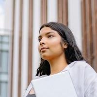 A young South Asian woman with long dark hair holding a laptop, standing outside a glass office building.