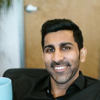 A smiling South Asian man in a black shirt holding a pale blue mug, seated by an office window.