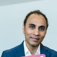 A South Asian man in a blue suit holding a pink folder, looking at the camera in an office.