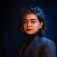 A South Asian woman with short dark hair in a dark blazer and turtleneck, lit against a near-black background.