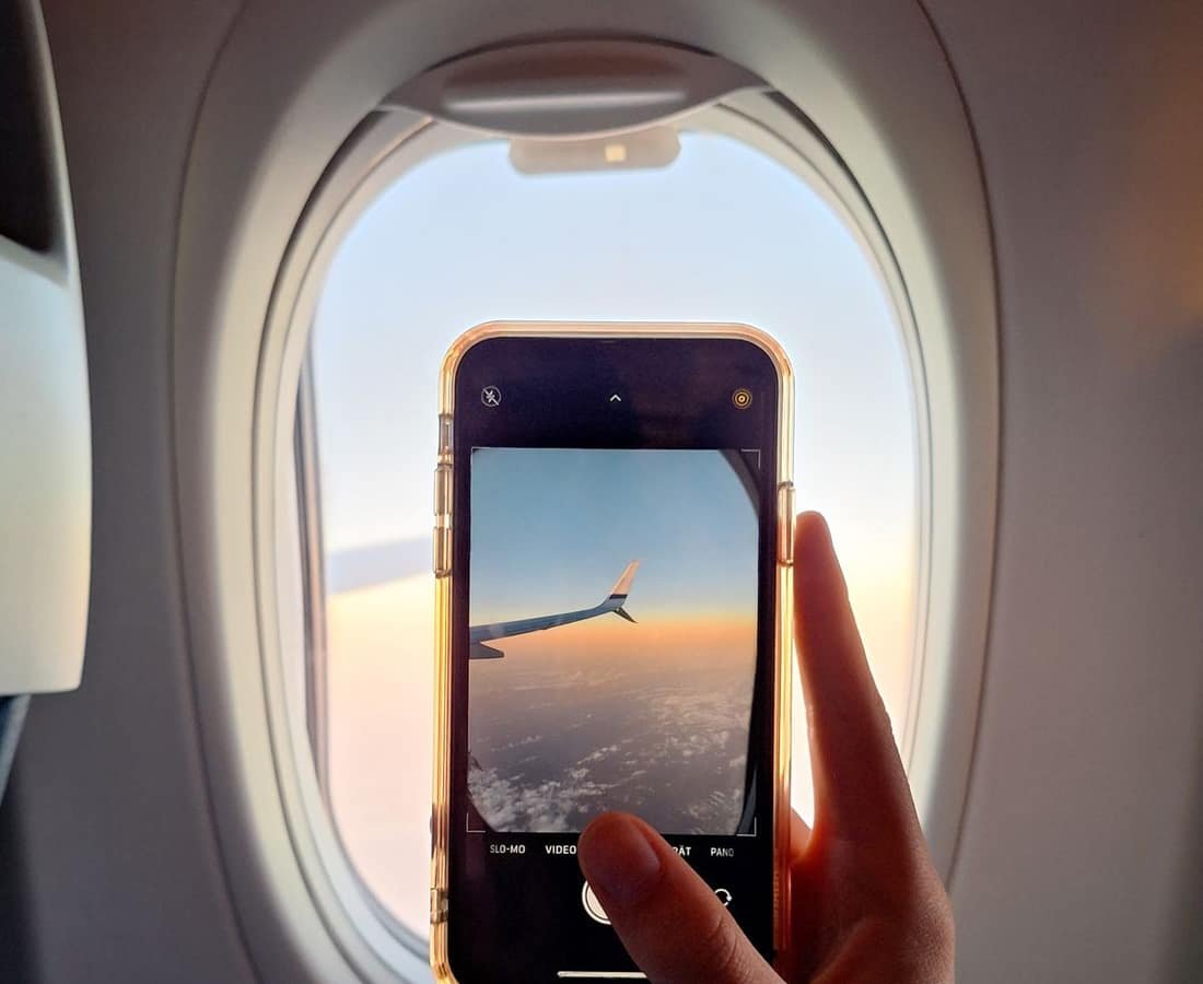 A hand holding a smartphone up to an aircraft window, the screen framing the plane's wing above a golden sunrise horizon and clouds.