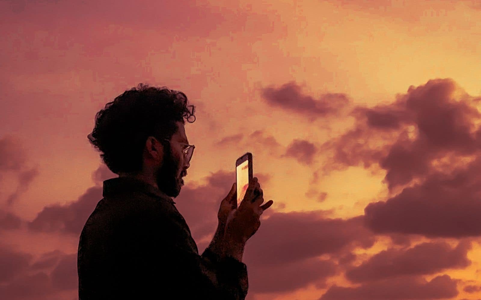 The silhouette of a curly-haired man holding up a smartphone, its screen glowing, against a warm pink and orange sunset sky over the sea.